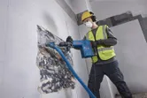 Eine Person, die Schutzausrüstung trägt, benutzt einen Abbruchhammer, um Wandfliesen zu zerschlagen.