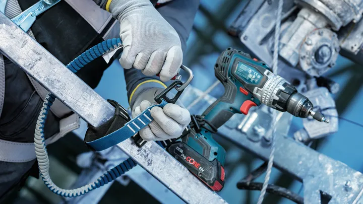 Eine Person mit Sicherheitsausrüstung befestigt ein Verbindungsmittel in der Nähe einer Akkubohrmaschine an einer Metallstruktur.