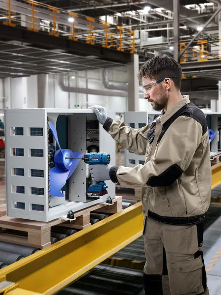 Eine Person, die Schutzausrüstung trägt, montiert mit einer Bohrmaschine einen Industrielüfter.