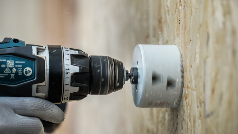 Eine Person, die Schutzausrüstung trägt, bohrt mit einem Elektrowerkzeug ein kreisrundes Loch in Holz.