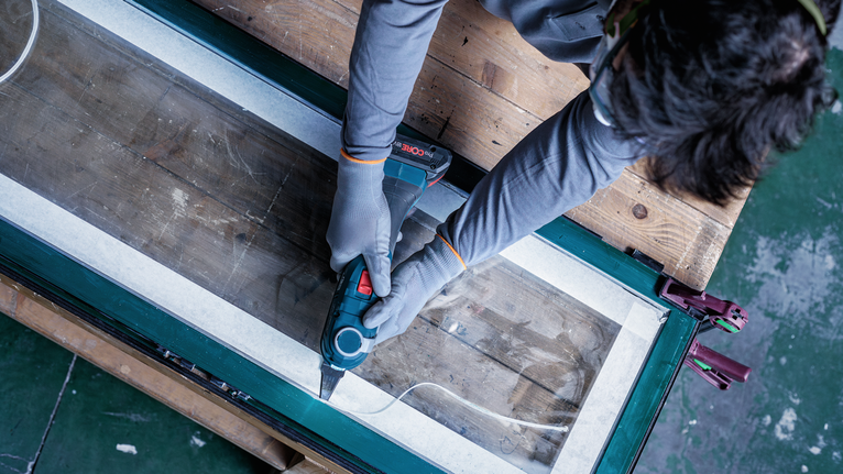 Eine Person, die Schutzausrüstung trägt, schneidet mit einem Elektrowerkzeug entlang eines abgeklebten Fensterrahmens.