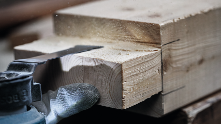 Ein Elektrowerkzeug schneidet durch einen dicken Holzbalken, Holzstaub wirbelt auf.