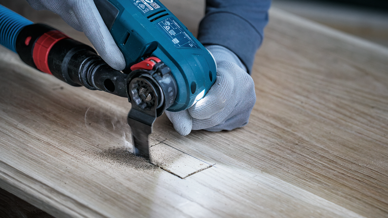 Eine Person, die Schutzausrüstung trägt, schneidet mit einem Elektrowerkzeug eine quadratische Öffnung in Holz.