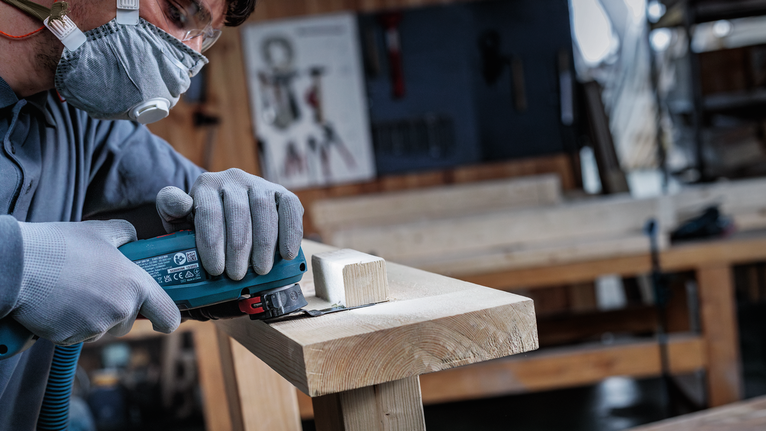Eine Person, die Schutzausrüstung trägt, schneidet in einer Werkstatt Holz mit einem Elektrowerkzeug.