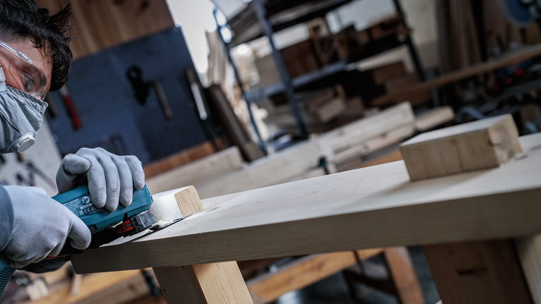 Eine Person, die Schutzausrüstung trägt, schleift in einer Werkstatt ein Holzbrett.