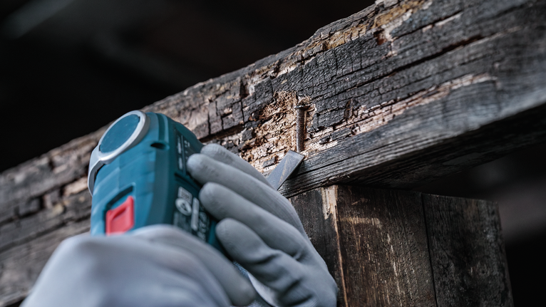 Eine Person, die Schutzausrüstung trägt, benutzt ein Elektrowerkzeug, um einen Nagel aus beschädigtem Holz zu entfernen.