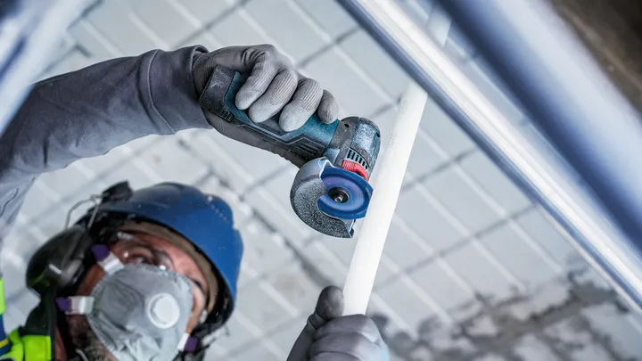 Eine Person mit Sicherheitsausrüstung schneidet mit einem kompakten Elektrowerkzeug ein weißes Rohr.