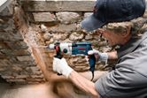 Eine Person mit Sicherheitsausrüstung bohrt in eine Steinmauer.