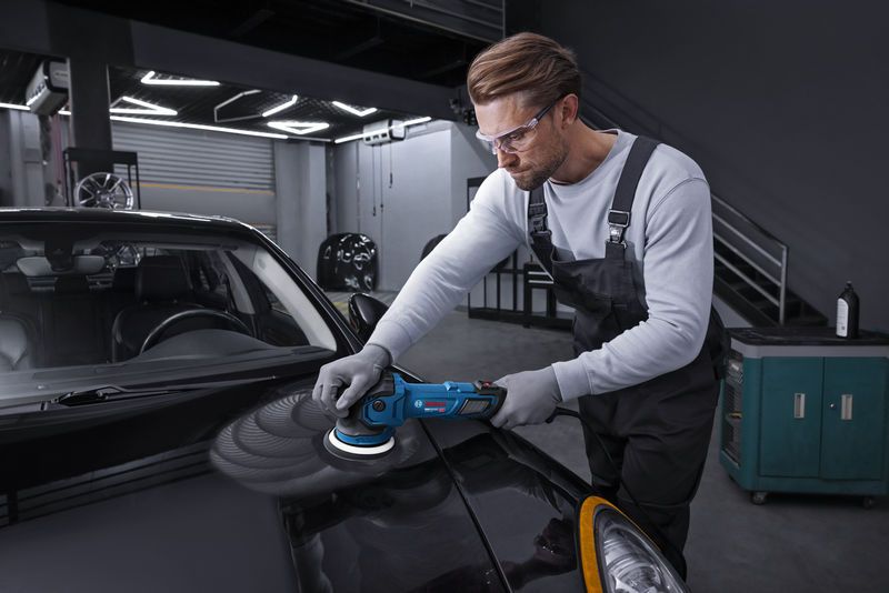 Eine Person in Schutzausrüstung poliert eine schwarze Automotorhaube mit einer Poliermaschine.
