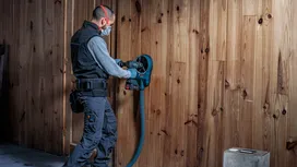 Eine Person, die Schutzausrüstung trägt, bohrt mit einem Elektrowerkzeug in eine Holzwand.