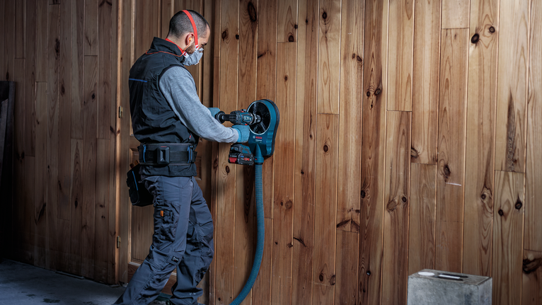 Eine Person, die Schutzausrüstung trägt, bohrt mit einem Elektrowerkzeug in eine Holzwand.