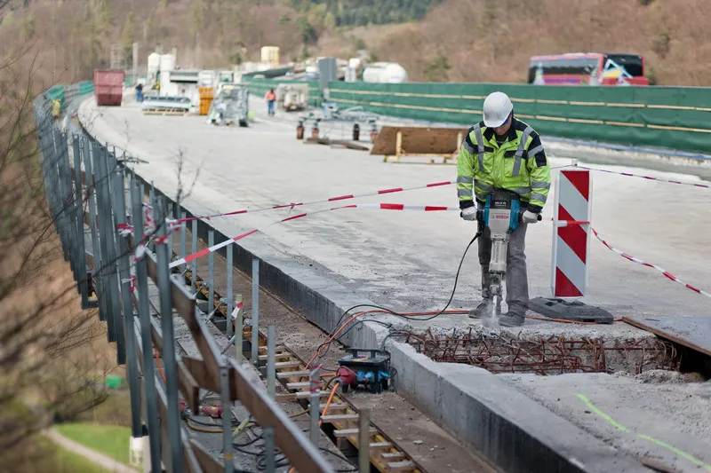 Eine Person mit Sicherheitsausrüstung bedient einen Brecher, um Beton auf einer Brücke abzureißen.