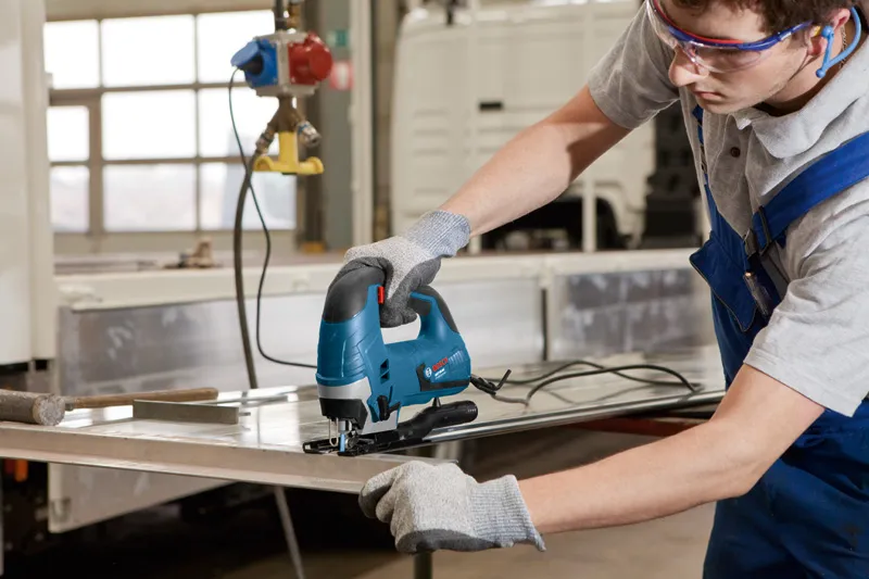Eine Person mit Schutzausrüstung schneidet mit einer Stichsäge ein Metallblech.