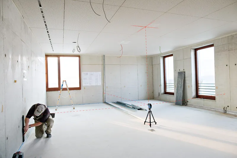Eine Person kniet, um eine Wand auszumessen, während ein Laser-Nivelliergerät Linien durch einen hellen Raum projiziert.