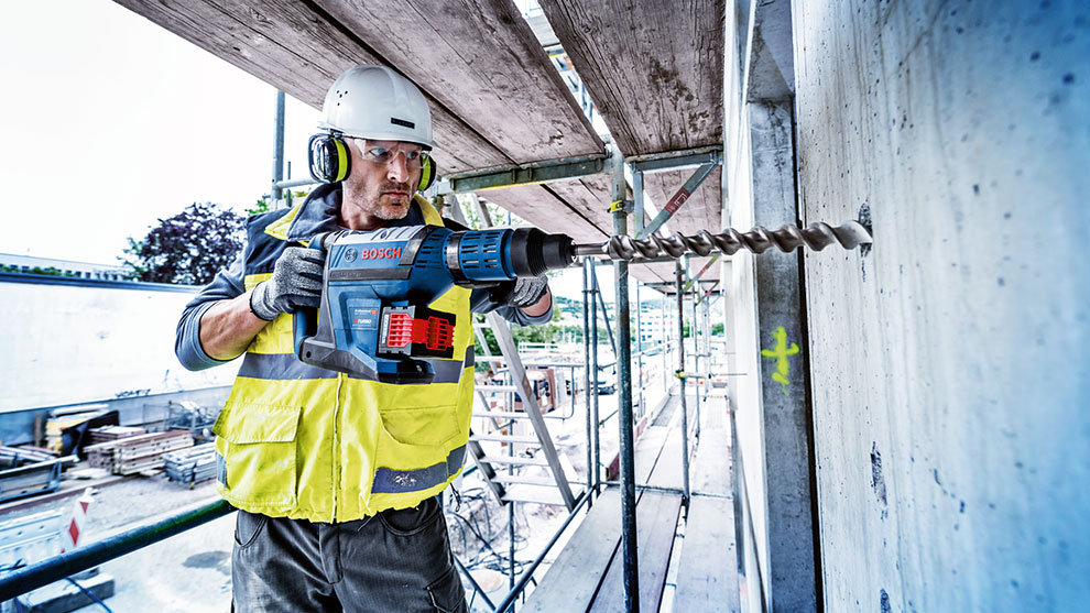Ein Arbeiter in gelber Weste bohrt mit einem blauen Bosch Werkzeug in eine Wand.