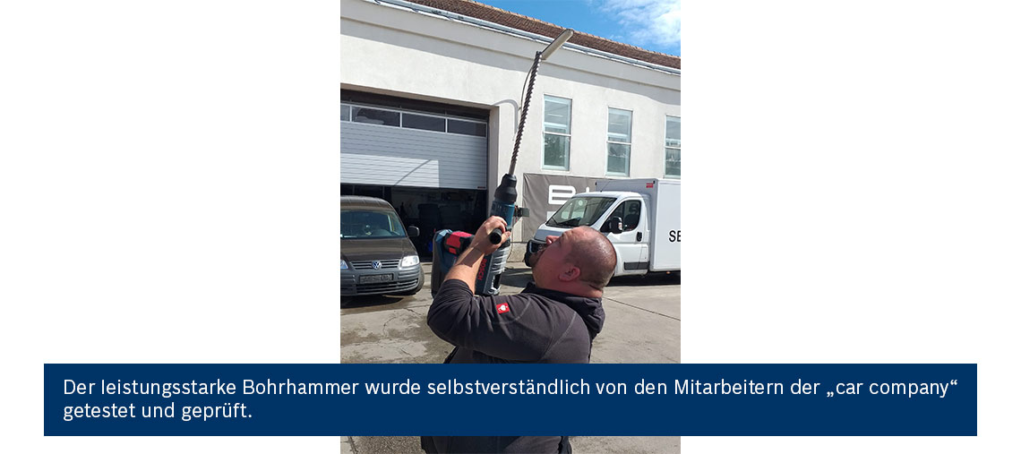 Ein Mann testet einen Bohrhammer im Freien, umgeben von Fahrzeugen und einem Gebäude.