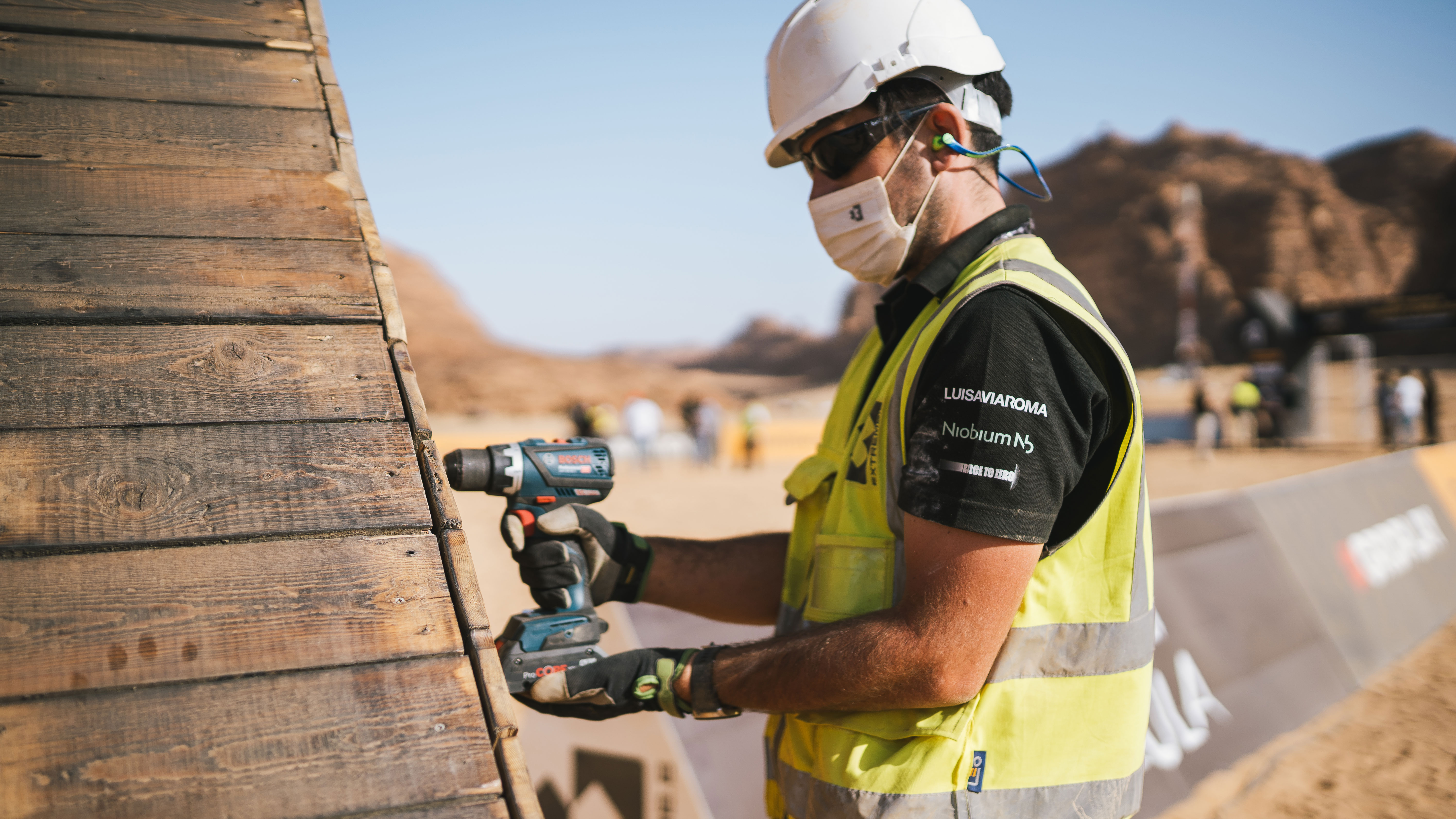Ein Arbeiter mit Bosch Akkuschrauber an einer Holzwand in einer Baustellenumgebung.