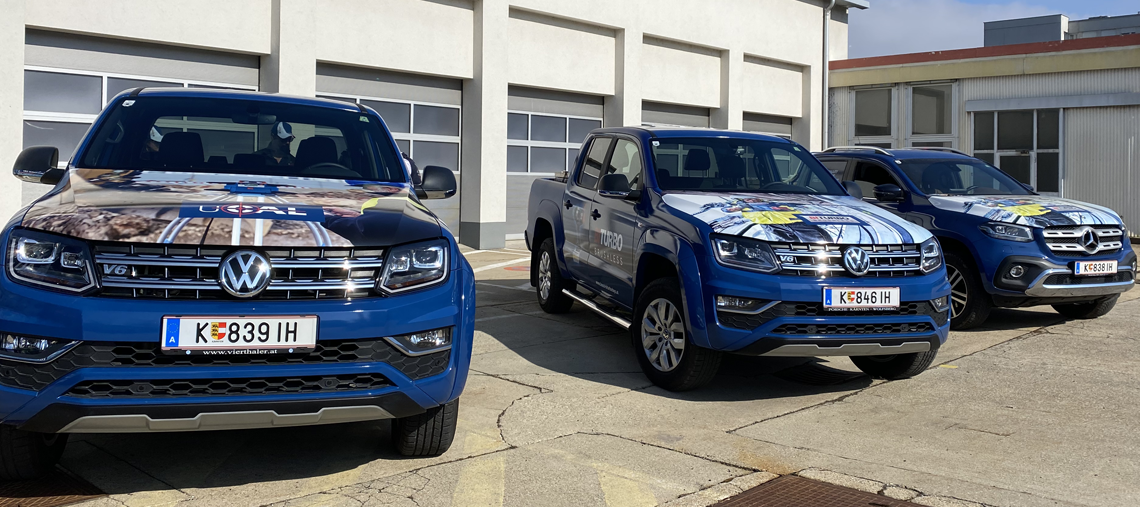 Drei Fahrzeuge stehen auf einem Parkplatz, blau mit Werbebeschriftungen.