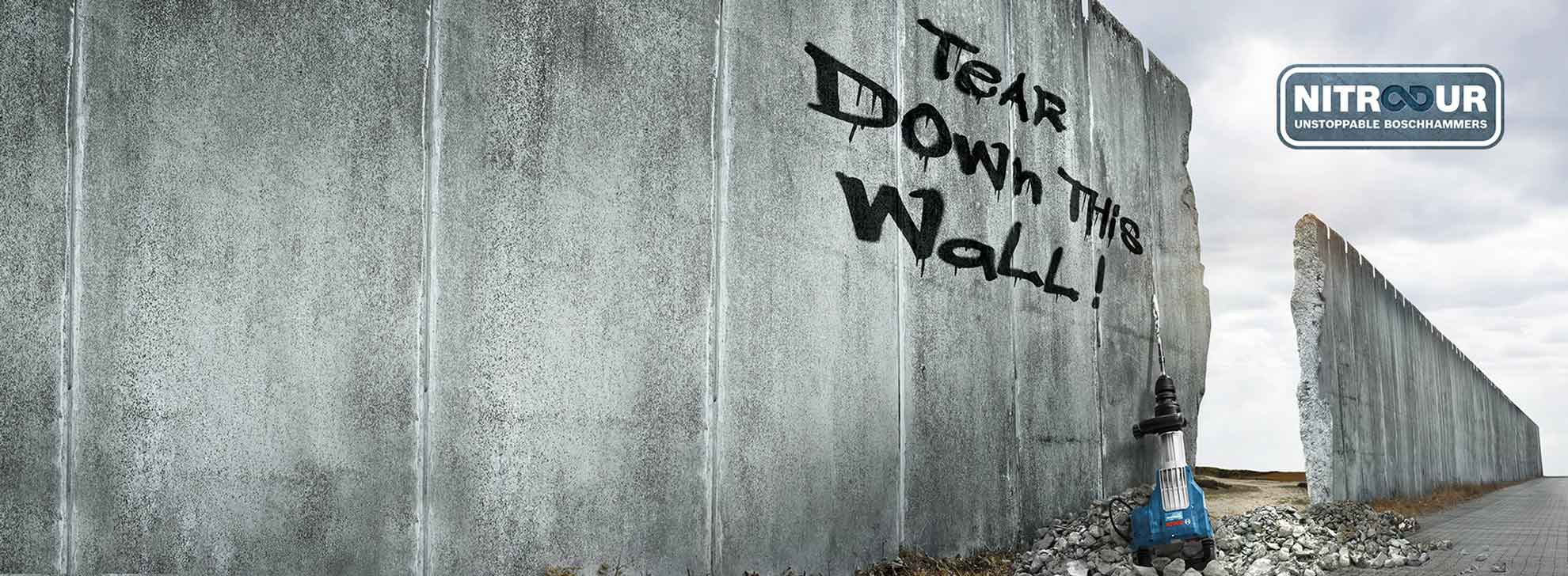 Ein blauer Bosch Hammer steht vor einer grauen Wand mit Graffiti: "Tear Down This Wall!"