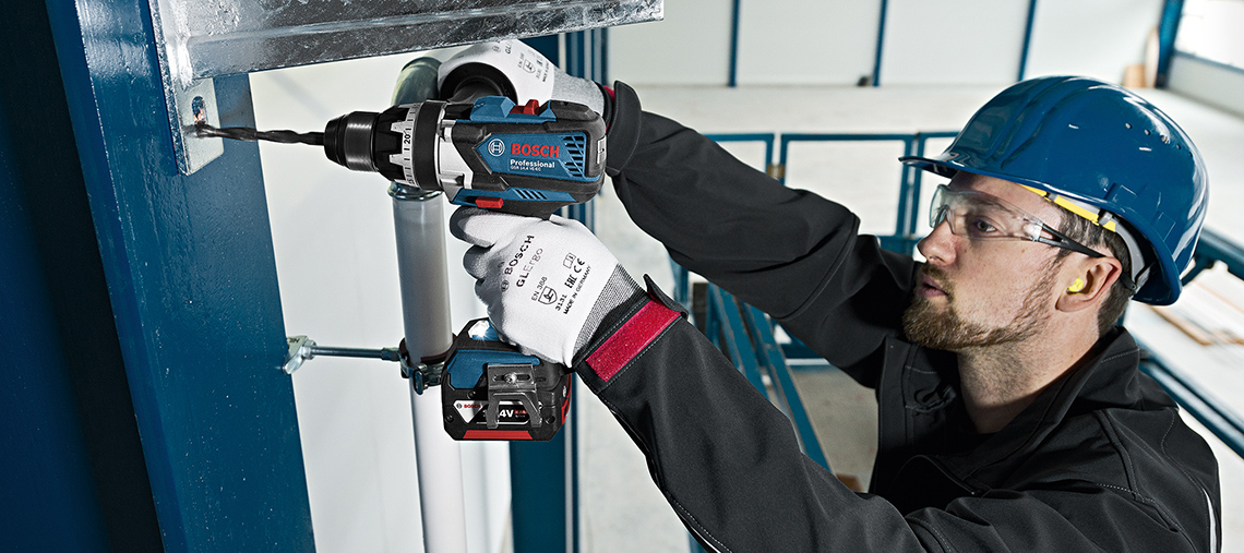 Ein Mann mit Helm bohrt mit einem blauen Bosch Professional Akku-Werkzeug in eine Wand.