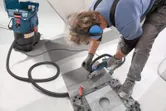 Une personne portant un équipement de sécurité coupe une tuile avec un outil électrique relié à un extracteur de poussière.