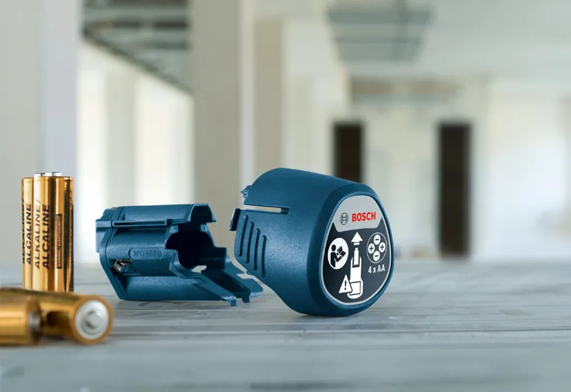 Accessoire adaptateur de batterie et quatre piles AA placés sur une surface en bois dans une pièce.