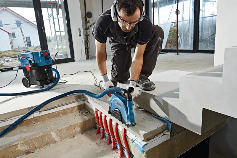 Une personne portant un équipement de sécurité utilise une rainureuse murale sur du béton au-dessus de tuyaux exposés.