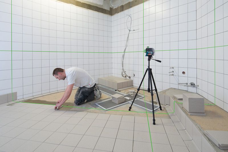 Personne installant des carreaux dans une salle de bain, à l'aide d'un outil de nivellement laser sur un trépied.