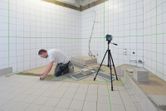 Personne installant des carreaux dans une salle de bain, à l'aide d'un outil de nivellement laser sur un trépied.