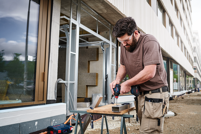 Un ouvrier portant un équipement de sécurité utilise une perceuse sans fil sur un cadre métallique sur un chantier de construction.
