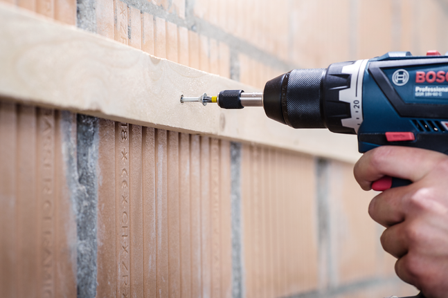 Personne utilisant une perceuse sans fil pour visser une vis dans une planche de bois fixée à un mur de briques.