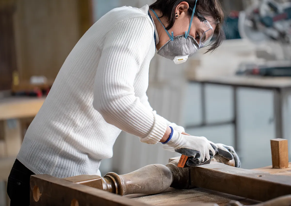 Une personne portant un équipement de sécurité ponce un pied de table en bois dans un atelier.
