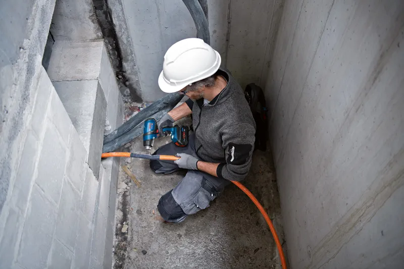 Une personne portant un équipement de sécurité perce un mur pour installer un conduit orange.