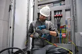 Une personne portant un équipement de sécurité utilise un pistolet thermique sans fil sur le câblage à l'intérieur d'un tableau électrique.