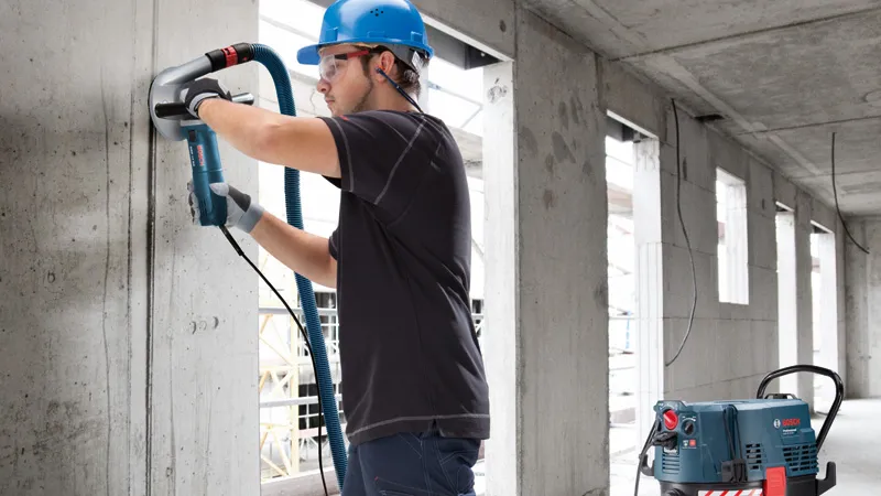 Une personne portant un équipement de sécurité ponce un mur en béton, un aspirateur à poussière se trouvant à proximité.