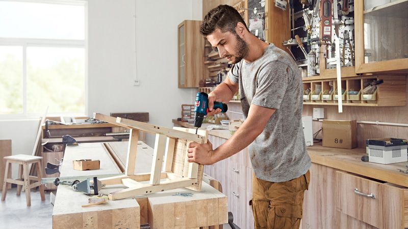 Une personne assemble une chaise en bois à l’aide d’une perceuse sans fil dans un atelier.