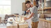 Une personne assemble une chaise en bois à l’aide d’une perceuse sans fil dans un atelier.