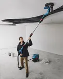 Une personne portant un équipement de sécurité ponce un plafond avec une ponceuse à cloison sèche dans un espace de rénovation.