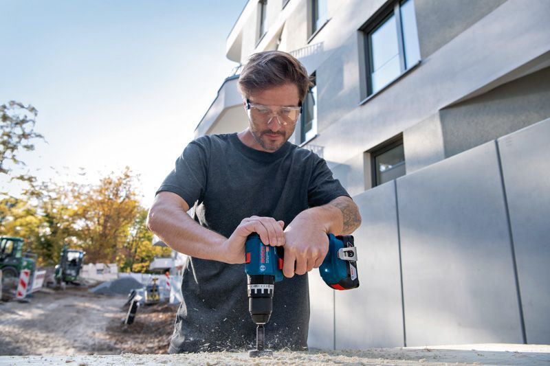 Une personne portant un équipement de sécurité utilise une perceuse sans fil pour percer un trou dans le bois à l'extérieur.