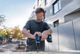 Une personne portant un équipement de sécurité utilise une perceuse sans fil pour percer un trou dans le bois à l'extérieur.