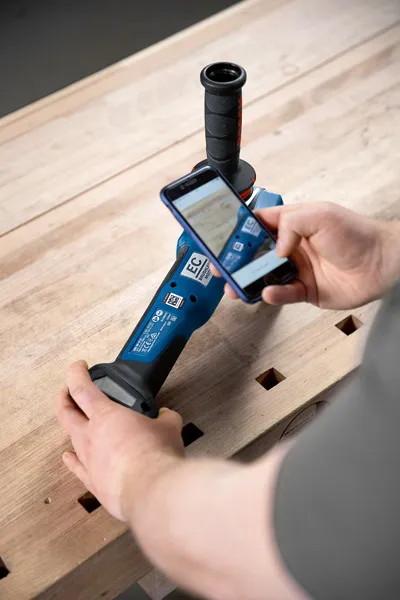 Une personne utilise un smartphone pour scanner l’étiquette d’un outil électrique sur un établi en bois.