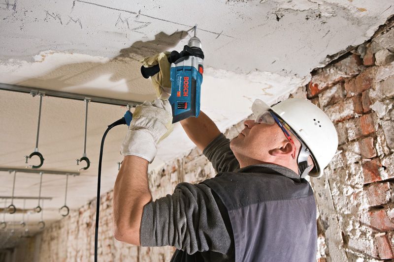 Une personne portant un équipement de sécurité perce un plafond en béton avec une perceuse à percussion.