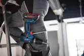 Une personne portant un équipement de sécurité se tient sur une échelle avec une perceuse combinée sans fil dans sa poche.