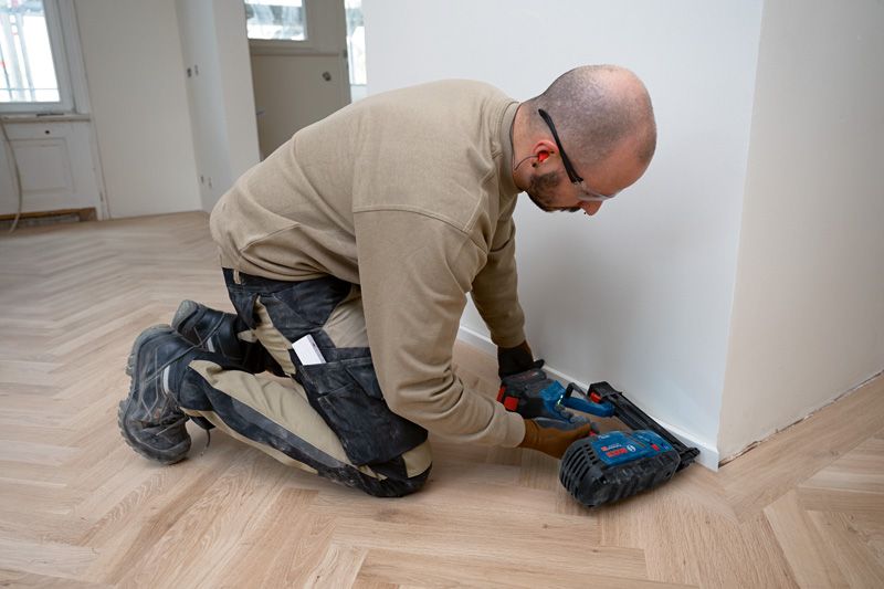 Une personne portant un équipement de sécurité utilise une cloueuse à bois sans fil pour installer des garnitures de plancher.