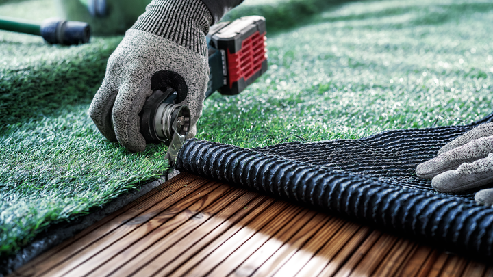 Une personne portant un équipement de sécurité coupe du gazon artificiel le long d'une terrasse en bois.