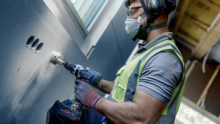 Une personne portant un équipement de sécurité perce un trou dans un mur à l’intérieur.