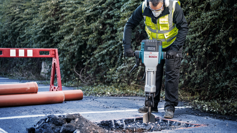Une personne portant un équipement de sécurité brise l'asphalte sur une route avec un marteau de démolition.