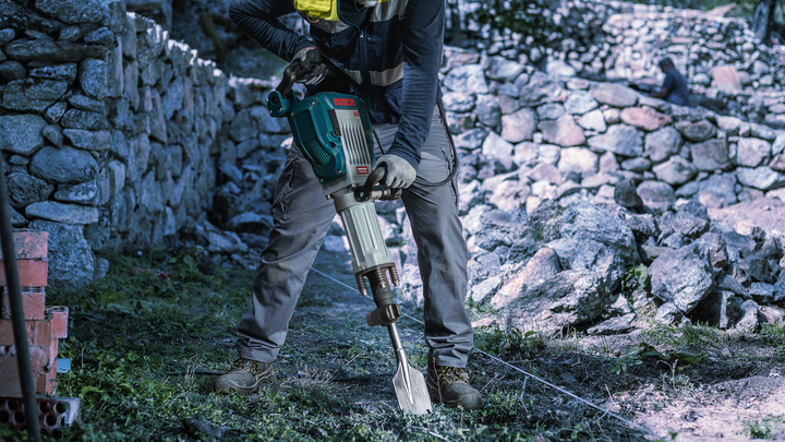 Une personne portant un équipement de sécurité utilise un marteau de démolition pour briser le sol près d'un mur de pierre.