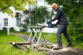 Une personne portant un équipement de sécurité utilise une tronçonneuse sans fil pour couper des bûches à l’extérieur.