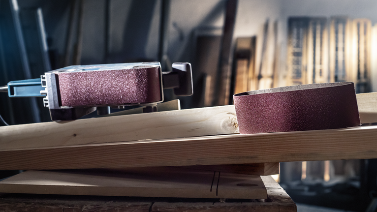Ponceuse à bande et bande abrasive posées sur une planche de bois dans un atelier.